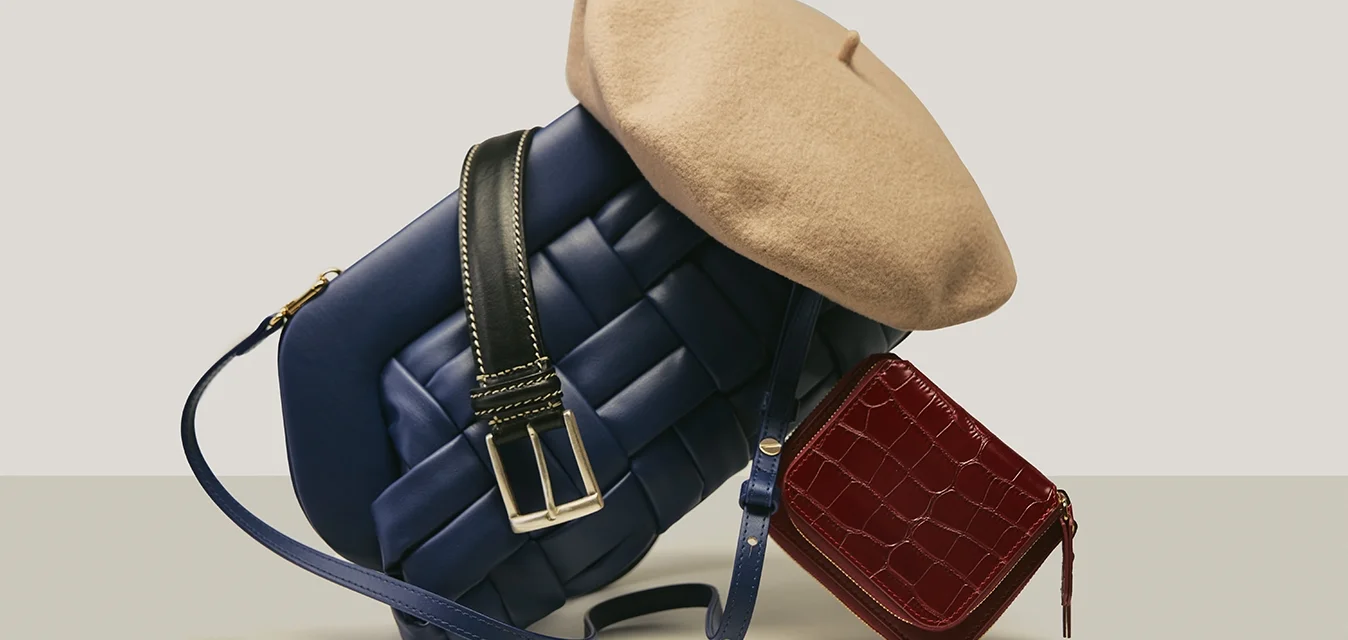 Borsa crossbody in pelle intrecciata blu con tracolla nera, dettagli in metallo argentato, berretto beige sopra e una piccola borsetta rossa con stampa coccodrillo.