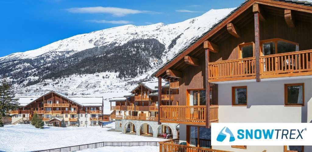 Snow-covered mountain ski resort with wooden alpine chalets and balconies under blue sky, SnowTrex logo visible