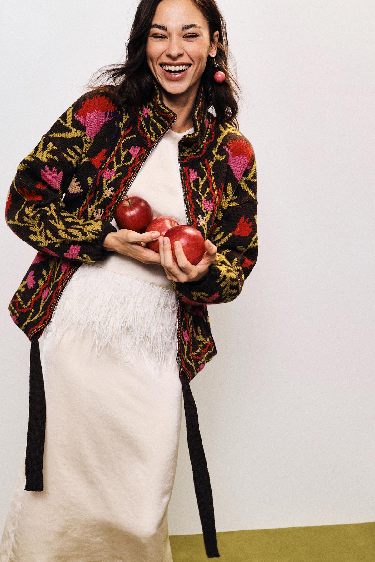 Smiling woman in floral knit jacket and white dress holding three red apples