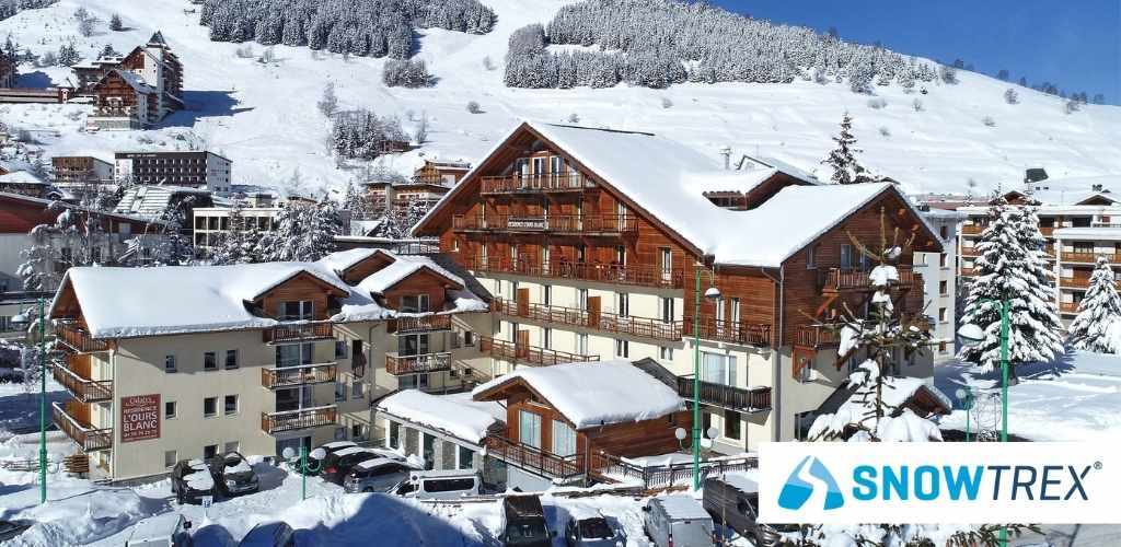 Snow-covered alpine ski resort with wooden chalets and car park at the base; SnowTrex logo visible