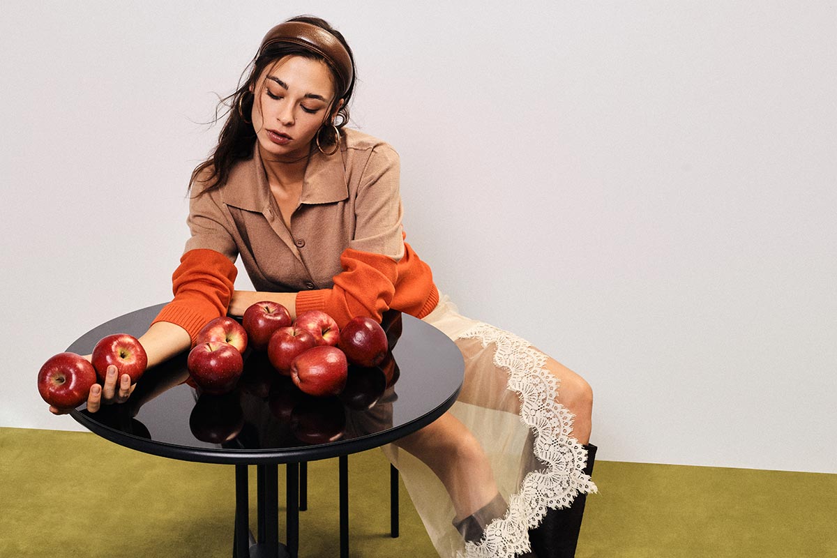 Young woman in beige and orange top with lace skirt seated at a black round table arranging red apples