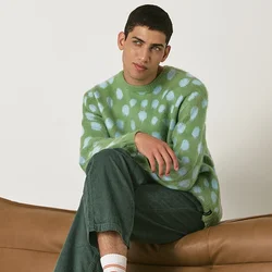 Maglione di maglia a pois verde e blu, pantaloni di velluto a coste verdi, sneakers bianche e grigie con strisce verdi, seduto su un divano in pelle marrone.