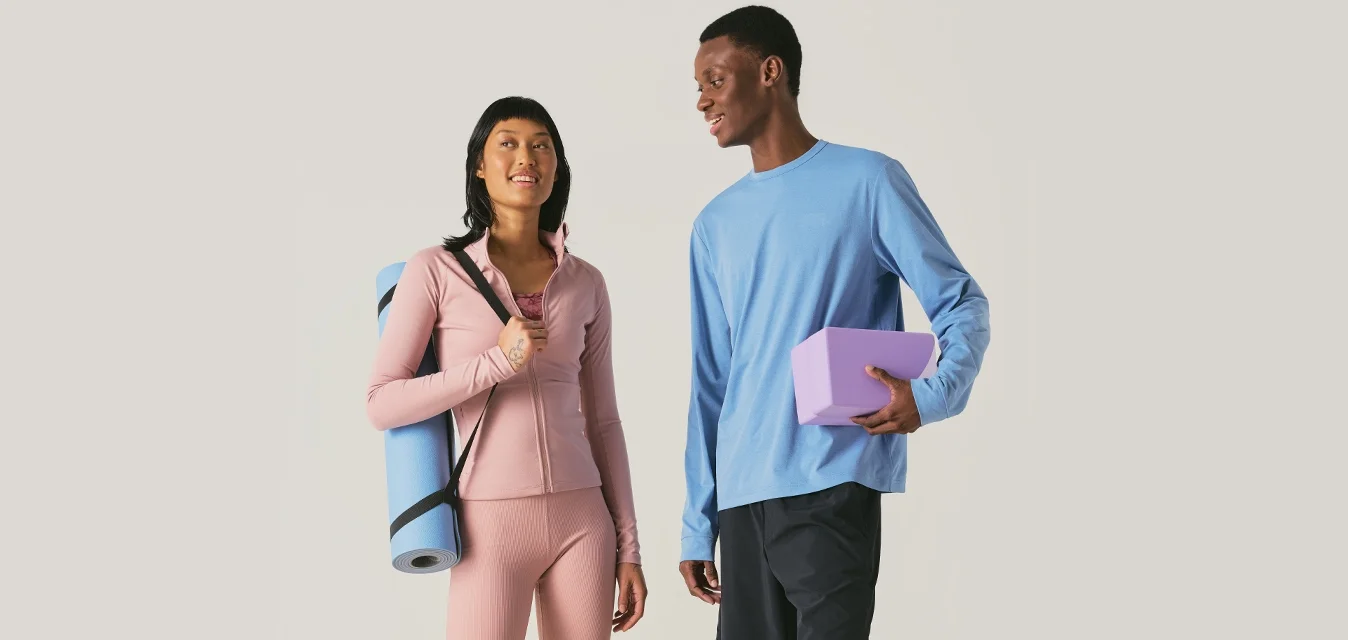 Donna con abbigliamento sportivo rosa tiene un tappetino da yoga blu; uomo con maglia blu e pantaloncini neri tiene un blocco da yoga viola, entrambi a piedi nudi e sorridenti.
