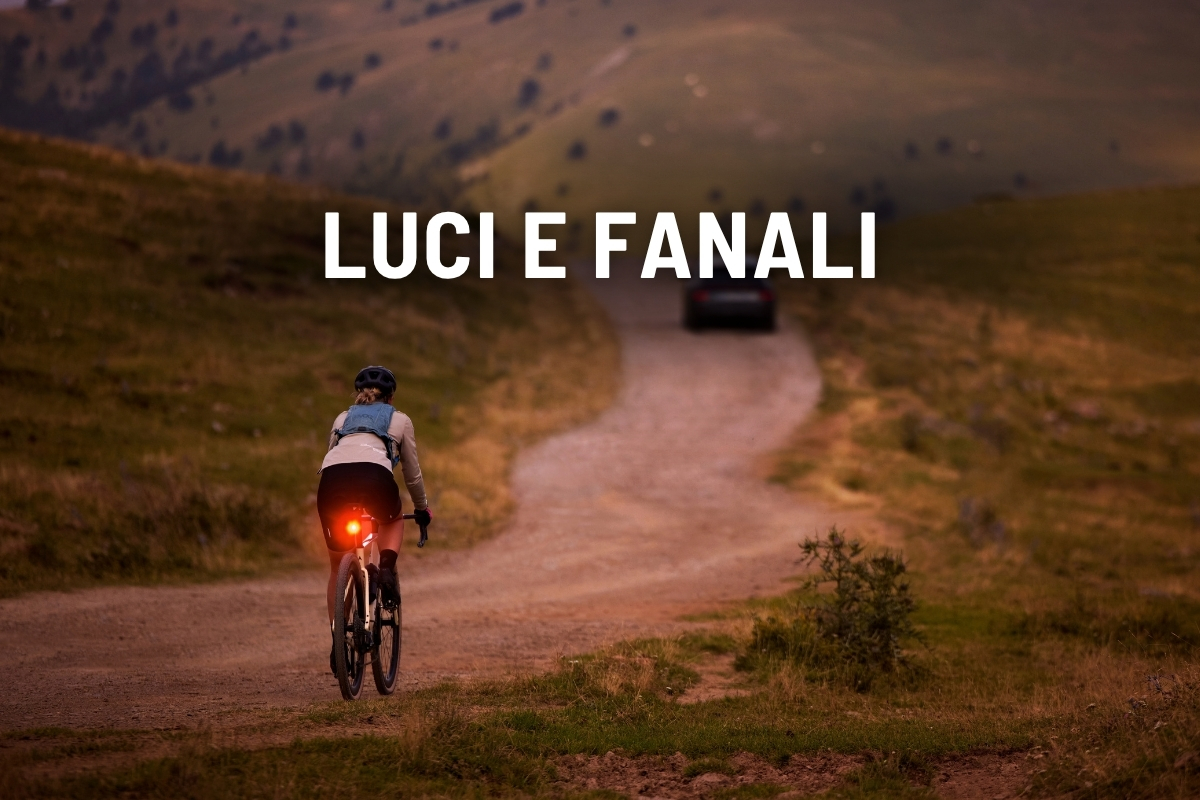 Cyclist riding on a rural dirt road at dusk with an illuminated red rear light, car ahead and 'LUCI E FANALI' text overlay