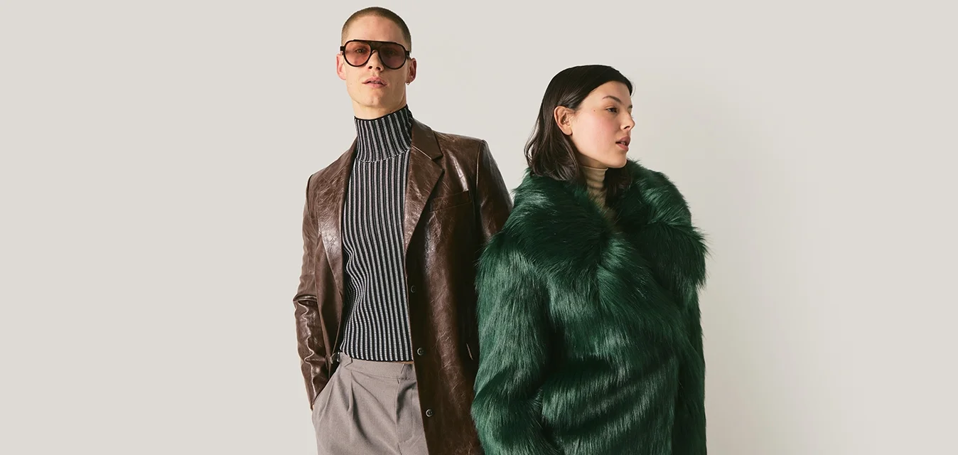 Due modelli in posa: uno con un cappotto di pelle marrone, dolcevita a righe e occhiali da sole; l'altro con un cappotto di pelliccia sintetica verde con le mani in tasca.