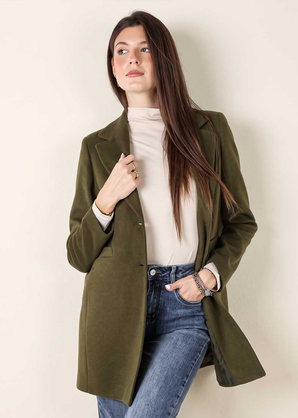 Woman wearing an olive-green coat over a beige turtleneck and blue jeans, long brown hair, hand in pocket, looking up against a pale wall