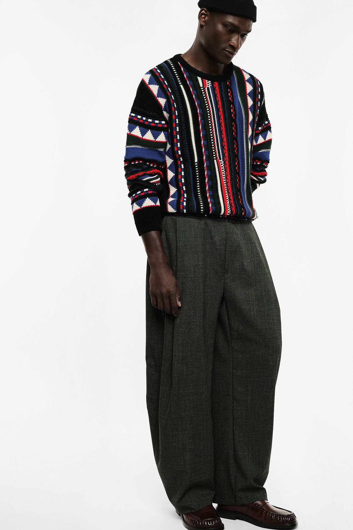 Black man wearing colourful patterned knit sweater, wide grey trousers and brown loafers, standing against white background