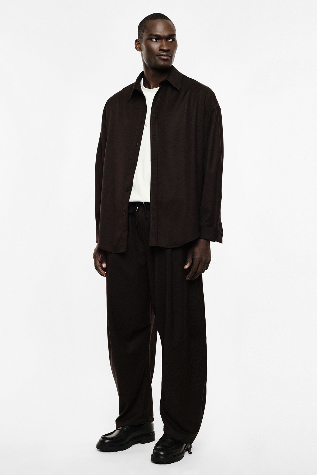 Man wearing a relaxed-fit dark brown shirt and wide trousers over a white T-shirt, black leather shoes