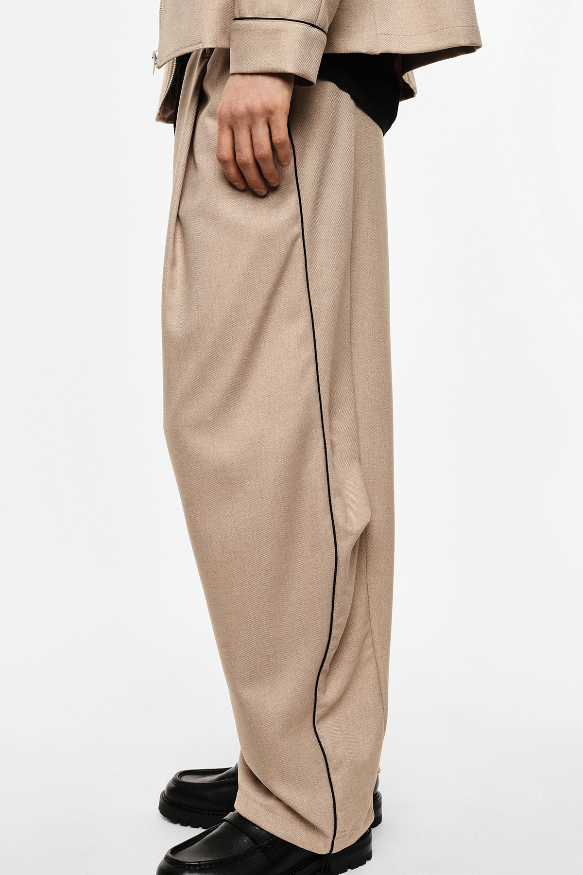 Side view of beige wide-leg trousers with black piping, matching jacket cuff and black chunky leather shoes, hand resting by side