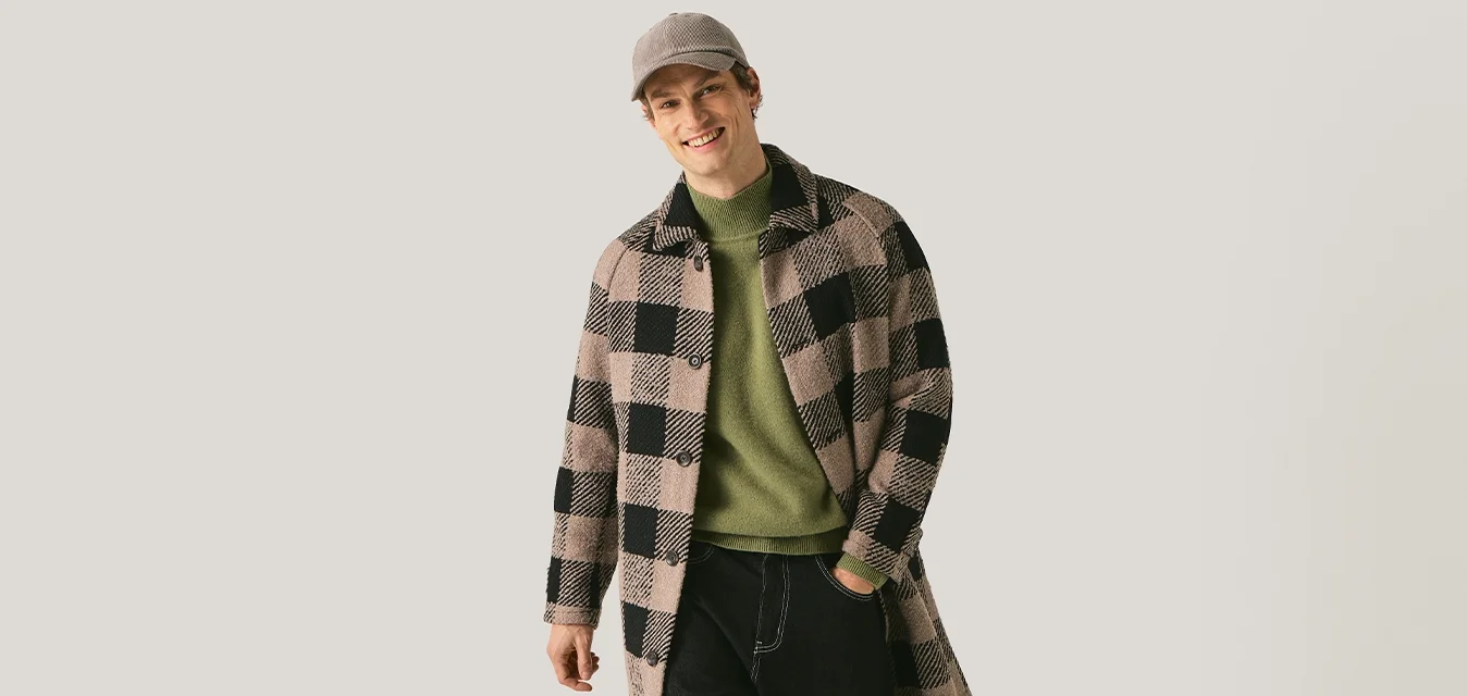 Uomo che cammina in avanti indossando un cappotto a quadri, maglione verde, pantaloni neri, scarpe marroni e un berretto a quadri abbinato su uno sfondo uniforme.