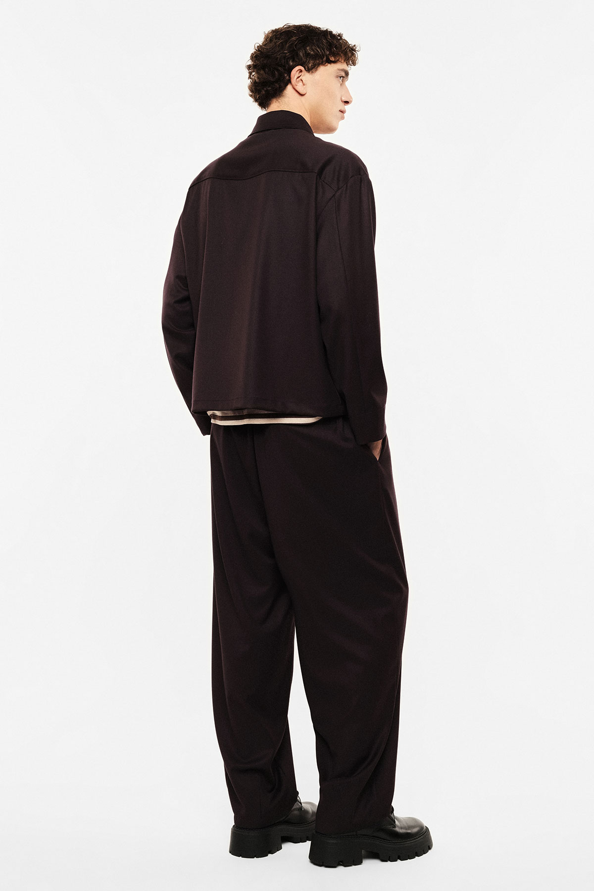 Rear view of a man modelling a dark relaxed-fit jacket, wide trousers and chunky black boots on a white studio background