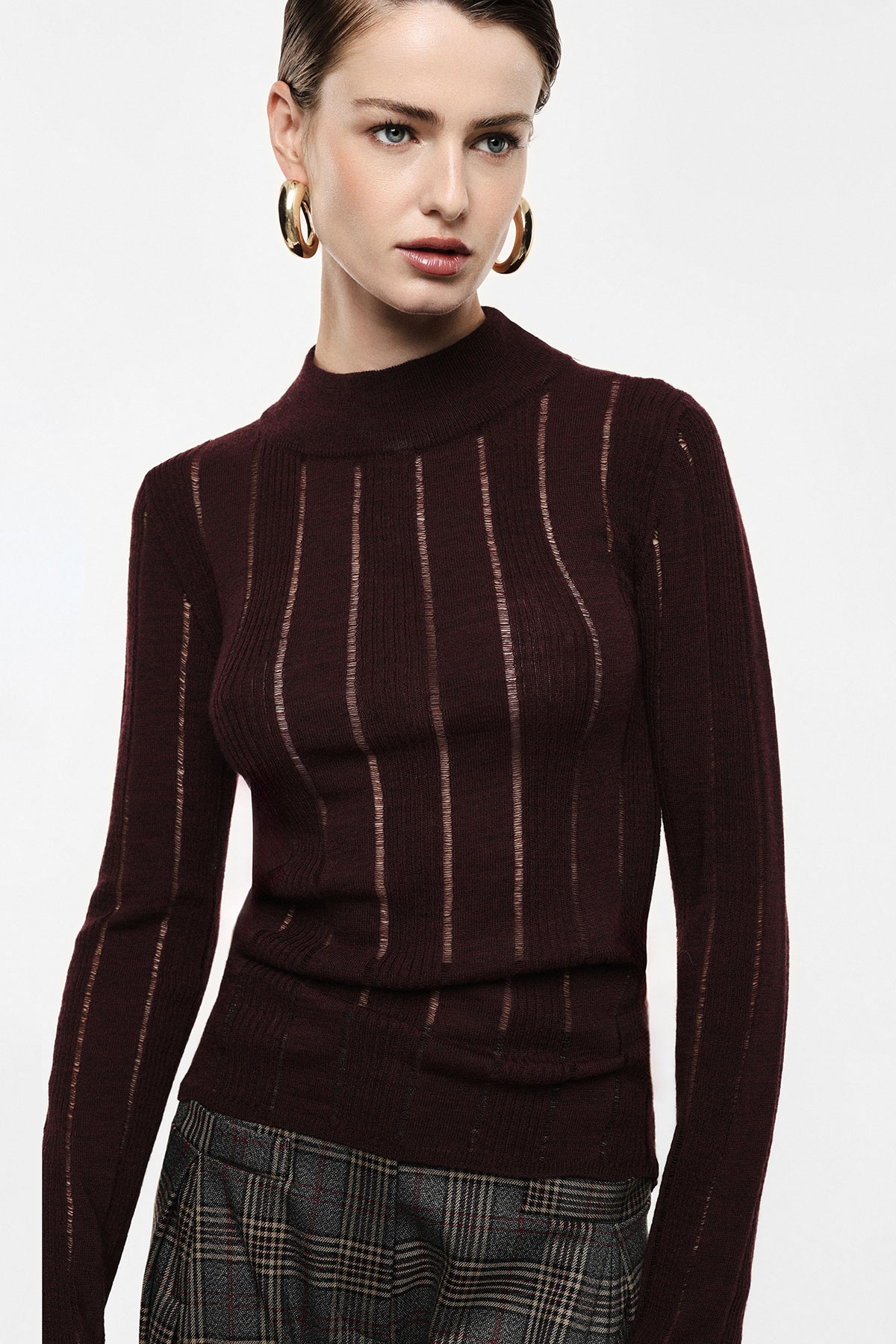 Studio portrait of female model wearing burgundy ribbed high-neck jumper, gold hoop earrings and grey checked trousers, looking right