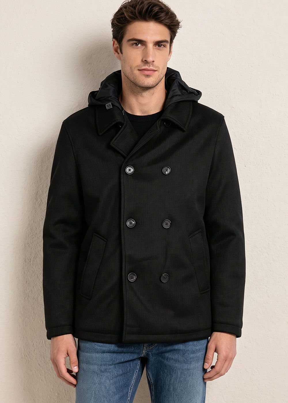 Man wearing a black double-breasted pea coat with a hood and blue jeans, standing against a neutral textured wall
