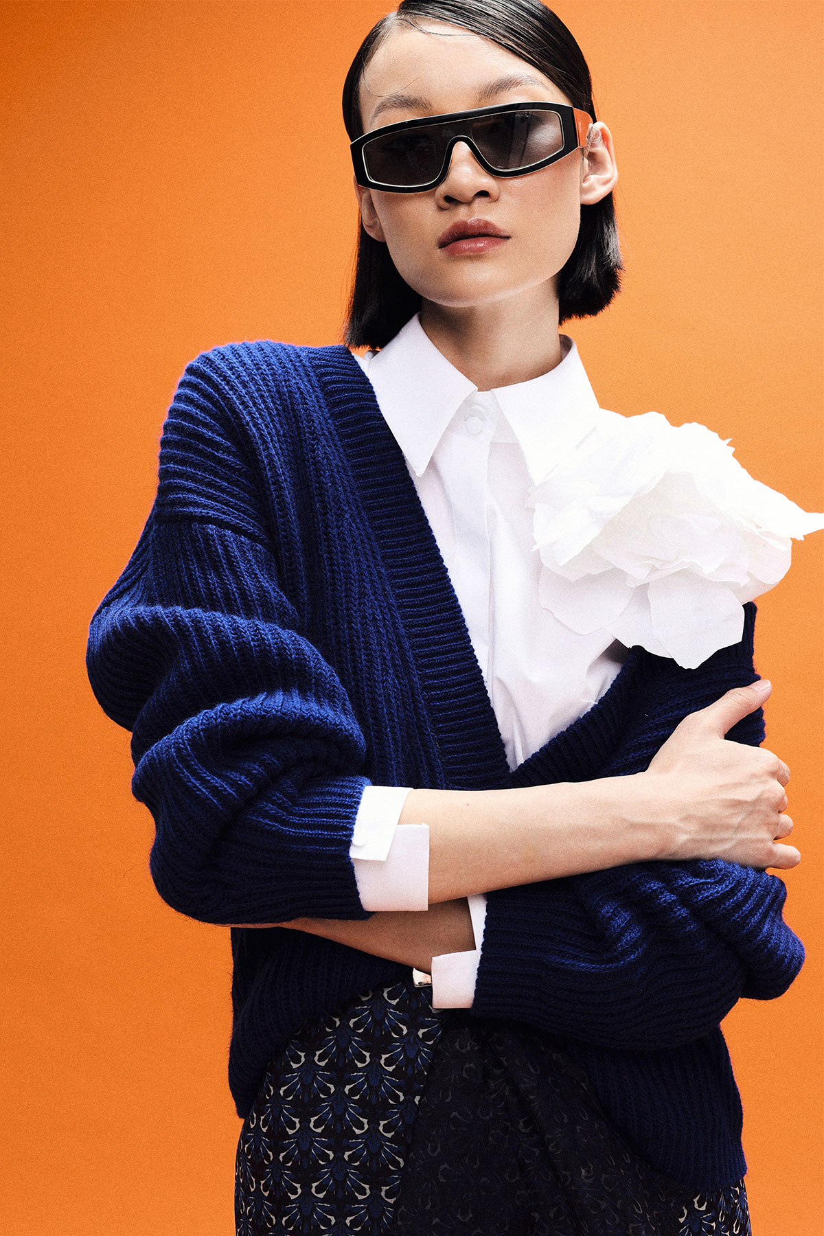 Fashion model wearing sunglasses, navy chunky cardigan and white blouse with fabric flower against orange background
