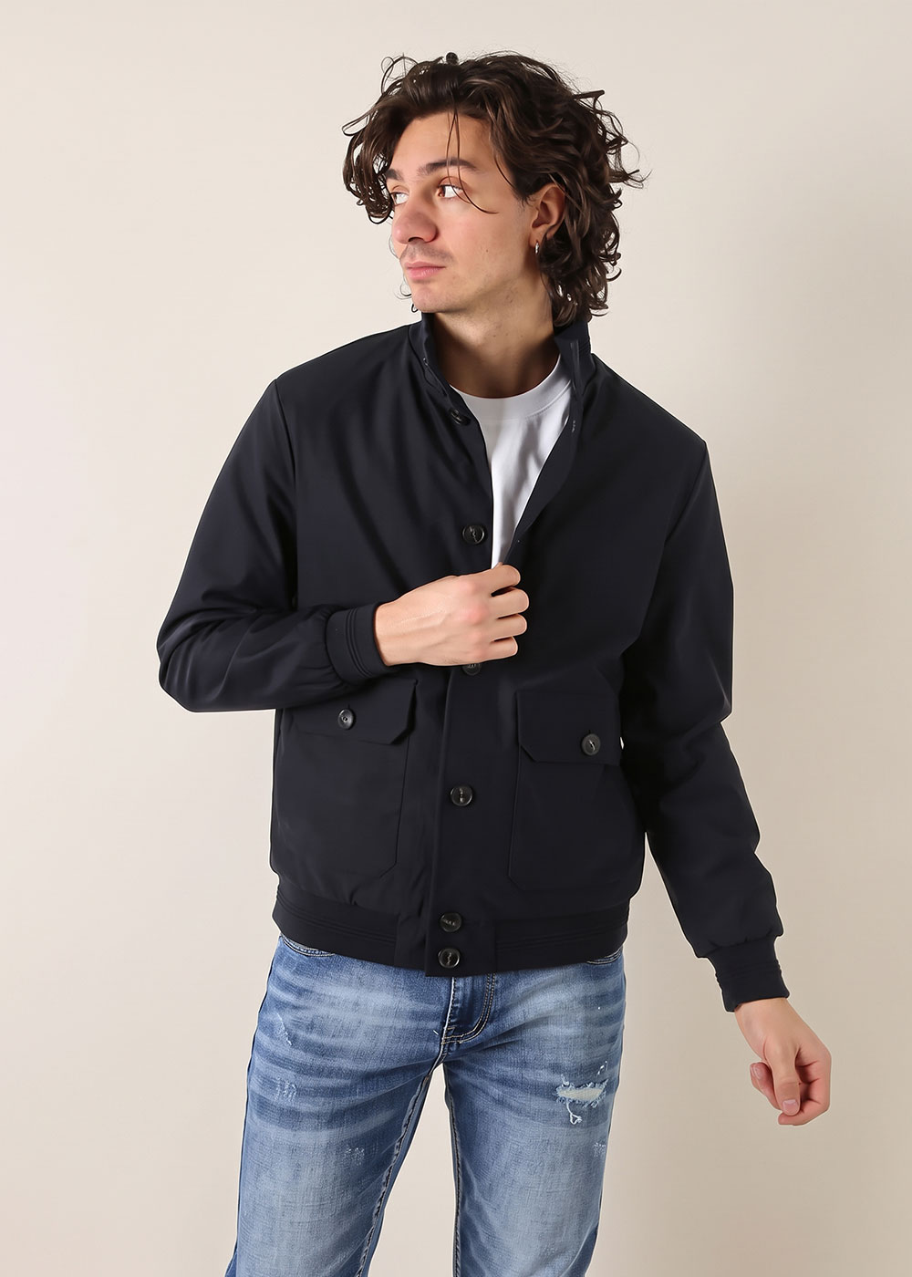 Young man looking left wearing a navy bomber jacket over a white T-shirt and ripped blue jeans, adjusting the jacket