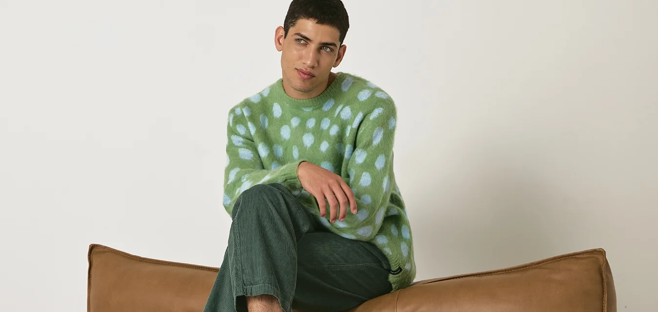 Maglione di maglia a pois verde e blu, pantaloni di velluto a coste verdi, sneakers bianche e grigie con strisce verdi, seduto su un divano in pelle marrone.