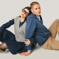 Due persone sedute a terra schiena contro schiena; una indossa una felpa con cappuccio blu e pantaloni kaki, l'altra un vestito di jeans, gilet grigio, stivali e occhiali da sole.