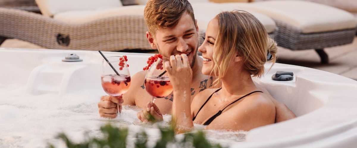 Young couple smiling in an outdoor hot tub, holding berry cocktails and enjoying a romantic spa moment