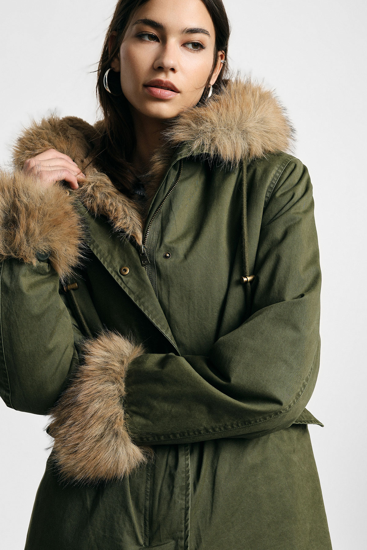 Young woman in olive-green parka with faux-fur hood and cuffs, arms crossed, wearing hoop earrings, looking to the side