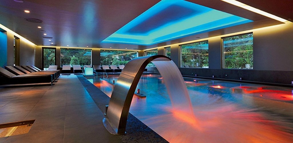 Indoor spa pool with illuminated waterfall, coloured LED lighting, lounge chairs and windows overlooking garden greenery