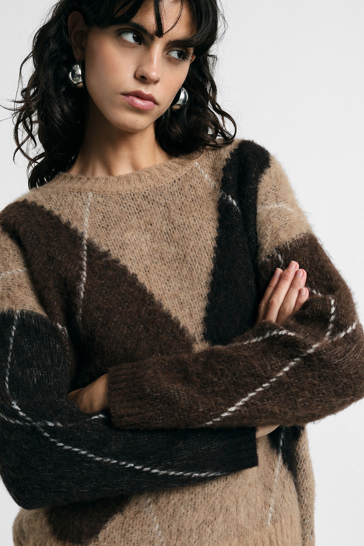 Woman wearing brown geometric mohair jumper with folded arms and silver hoop earrings, looking to the side