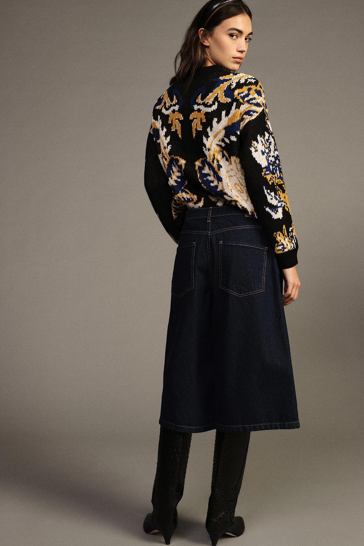 Young woman looking over her shoulder wearing a patterned black and gold knitted jumper, dark denim midi skirt and black knee-high boots on neutral background