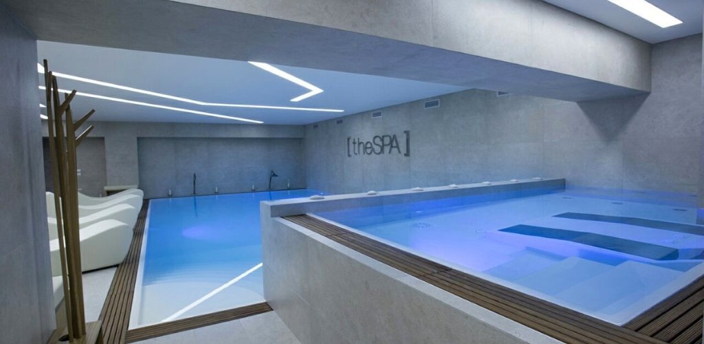 Modern indoor spa with illuminated blue pools, built-in lounger platforms and minimalist concrete interior