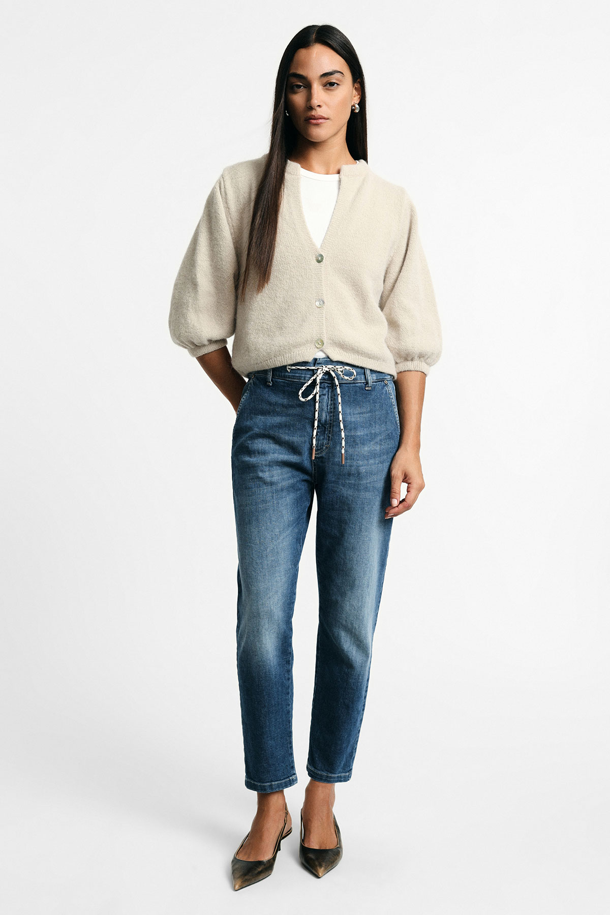 Woman in cream buttoned cardigan over a white top, high-rise blue drawstring jeans and pointed heels, full-length studio shot