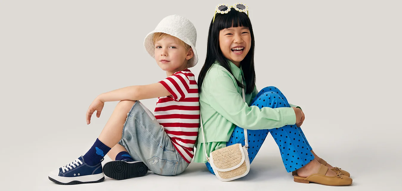 Due bambini sono seduti schiena contro schiena su un pavimento bianco semplice, uno indossa una camicia a righe e un cappello, l'altro pantaloni blu e una giacca verde sorridendo.