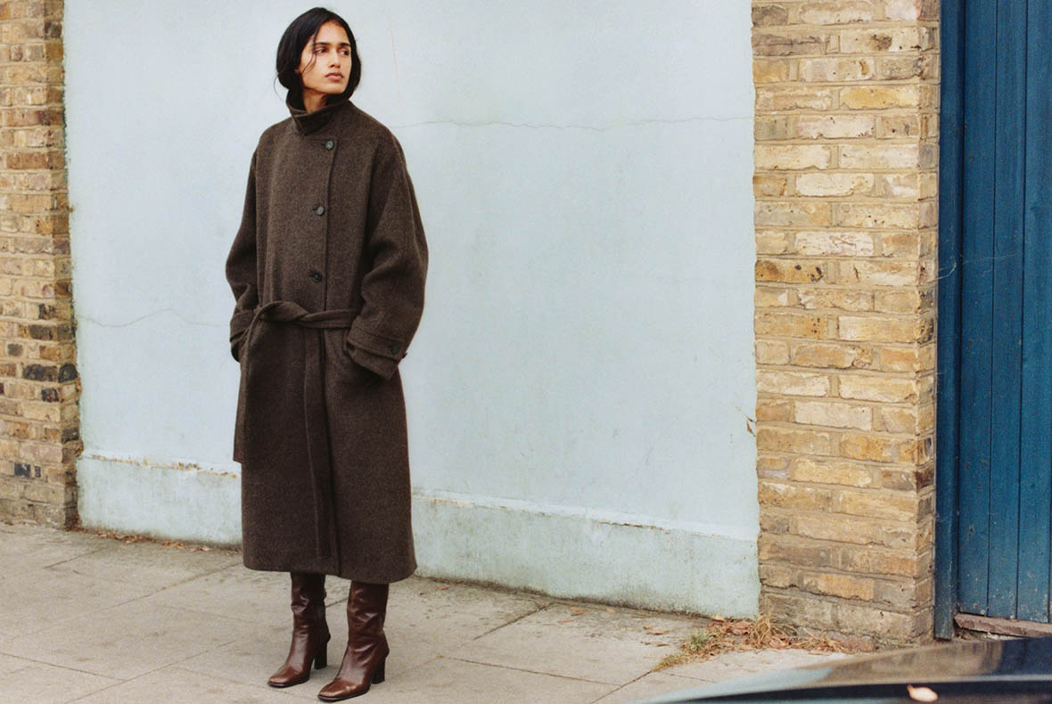 Person in long dark brown coat and leather heeled boots standing on pavement by pale wall and brick column.