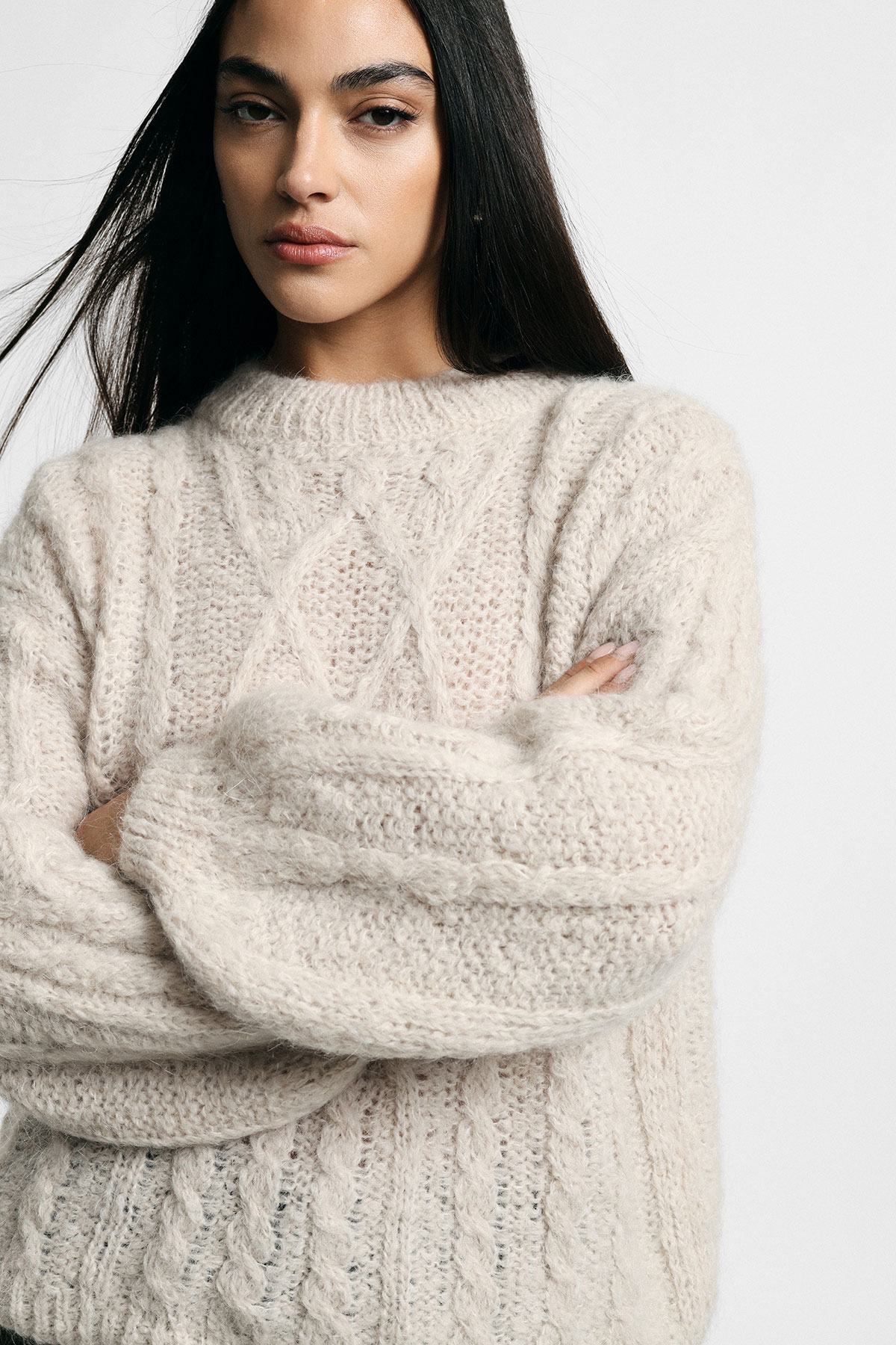 Close-up portrait of a woman with long dark hair wearing a cream cable-knit jumper, arms crossed with a neutral expression
