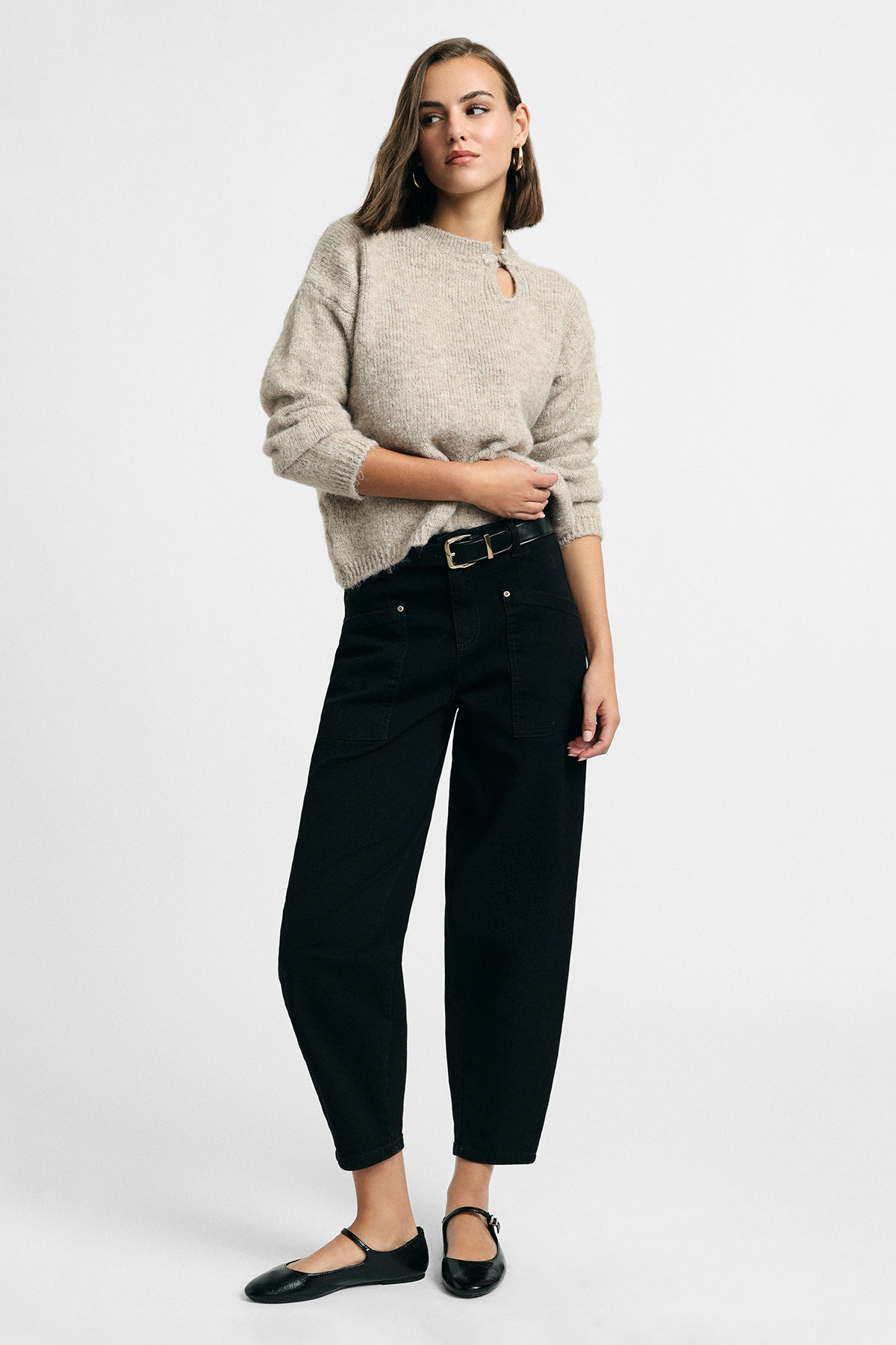 Woman in beige knit sweater with keyhole, high-waist black trousers and Mary Jane flats on neutral background