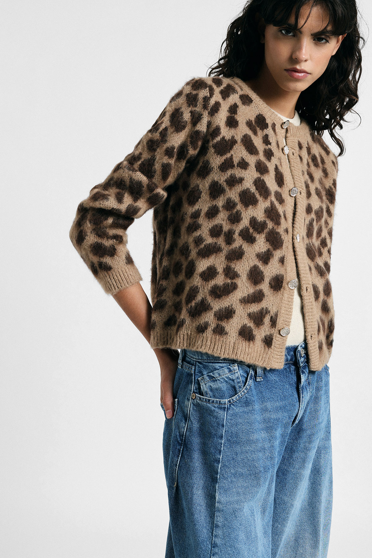 Woman wearing brown leopard-print fuzzy button-up cardigan over cream top with relaxed blue jeans, hands in pockets