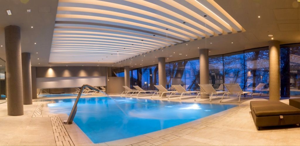 Indoor luxury hotel spa pool at dusk with glowing blue water, modern loungers, columns and floor-to-ceiling windows overlooking trees
