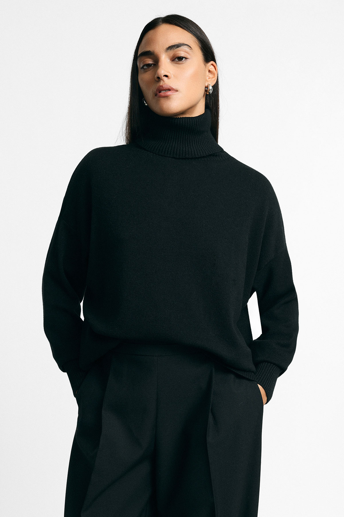 Woman in black roll-neck sweater and tailored black trousers, hands in pockets, standing against plain light background
