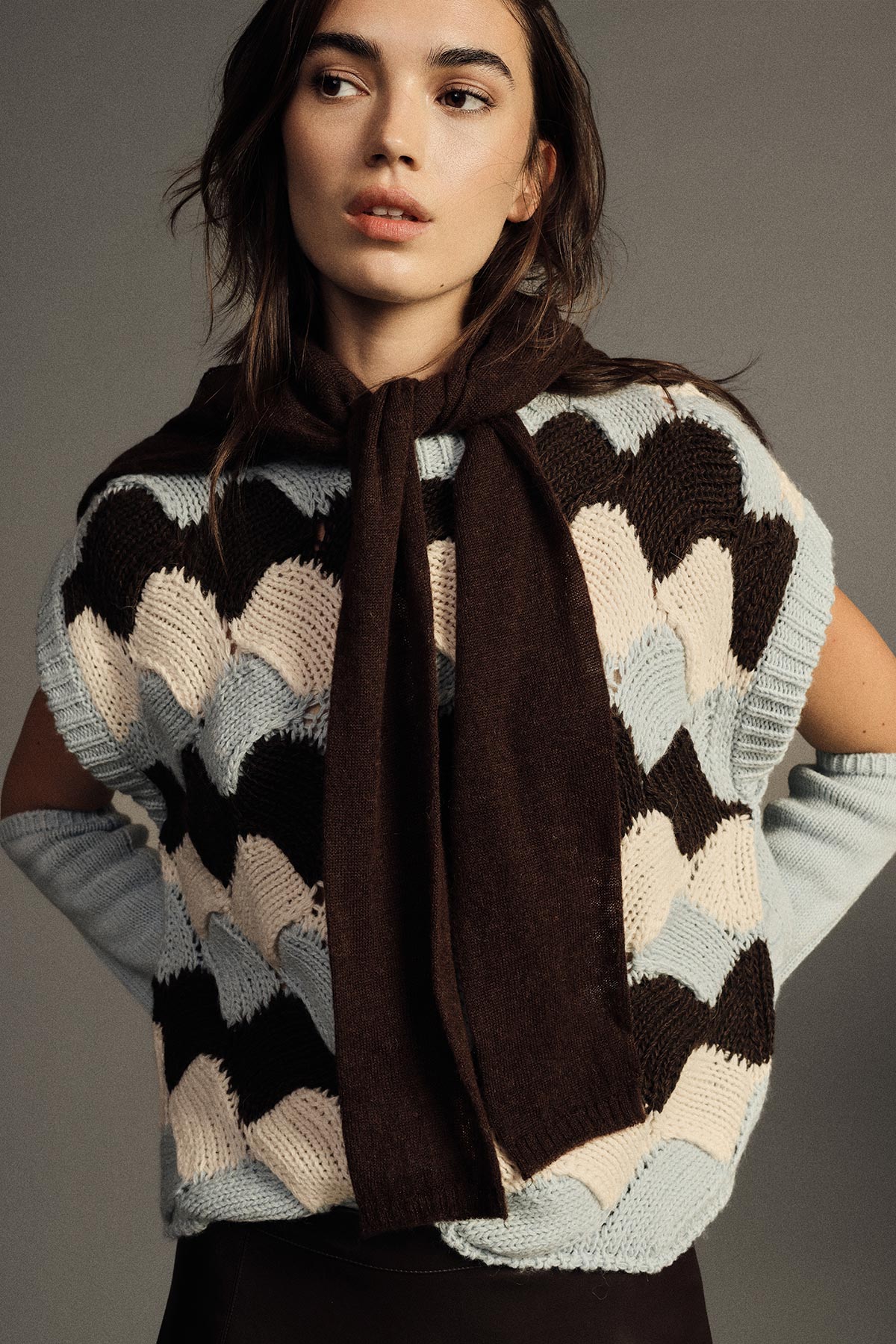 Woman modelling cream, blue and and brown scallop-knit sleeveless sweater with brown scarf in studio portrait