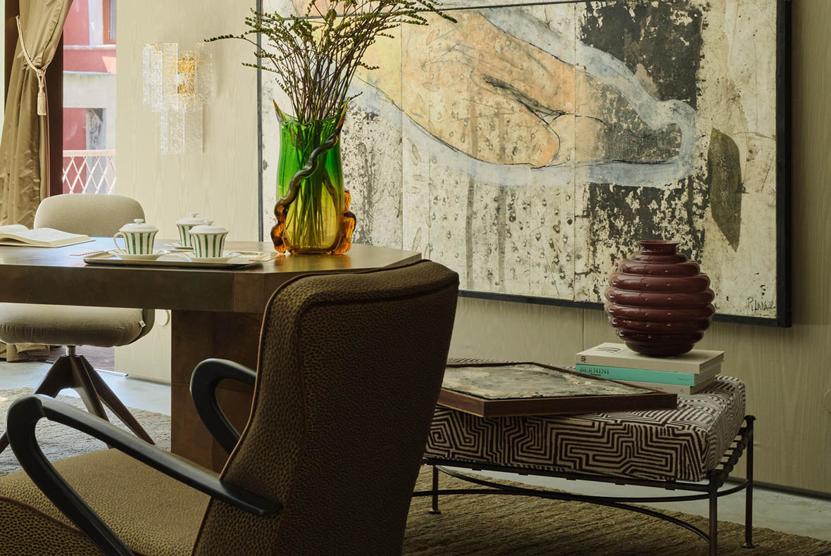 Stylish dining corner with wooden table, green glass vase of branches, abstract wall art, patterned ottoman and red ceramic vase
