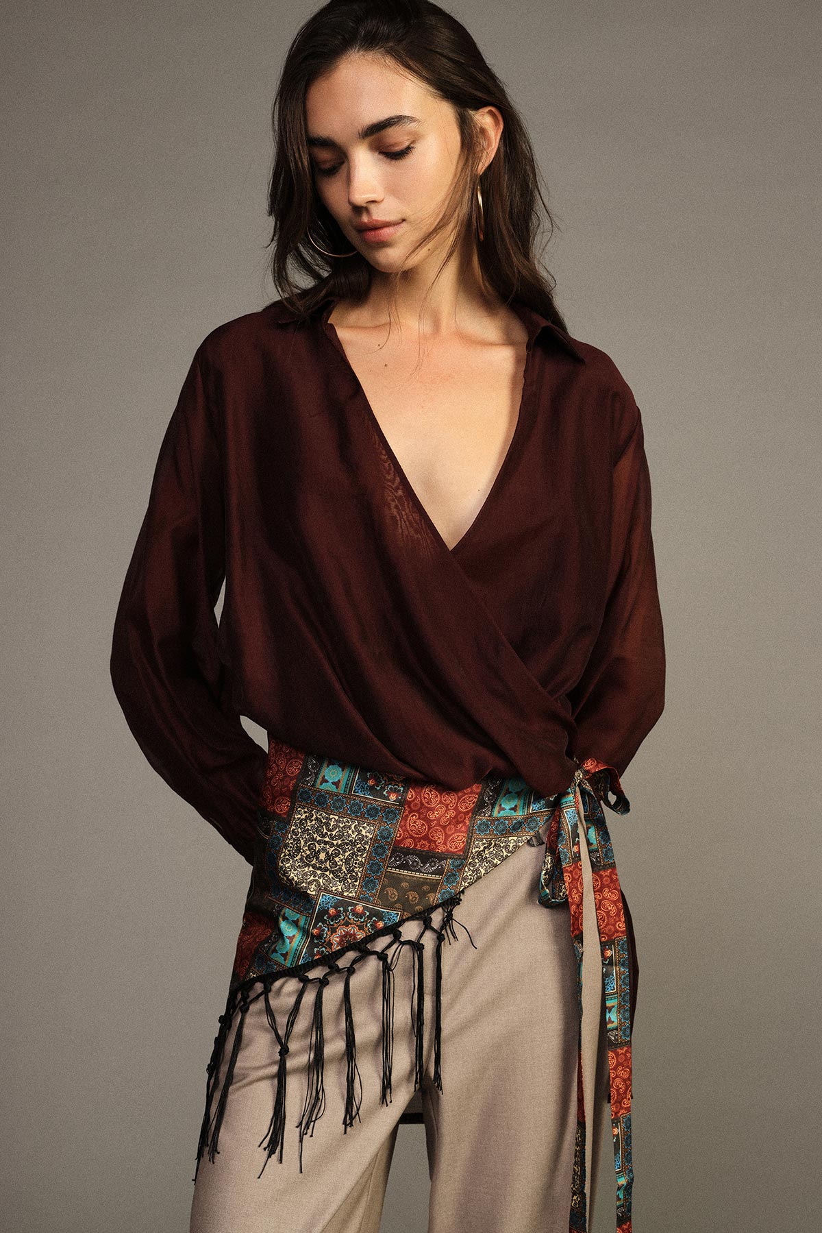 Woman wearing deep-burgundy wrap blouse and patterned fringed sash over beige trousers, looking down against a neutral background