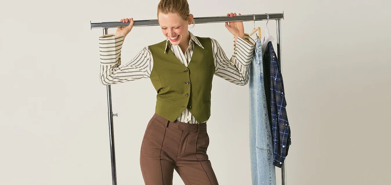 Donna con pantaloni marroni e gilet verde che tiene un attaccapanni con jeans e una camicia a quadri, in uno studio minimalista.