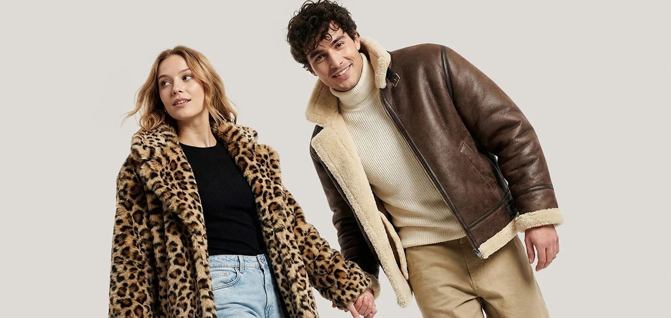 Un uomo e una donna che si tengono per mano, camminando in avanti; la donna indossa un cappotto con fantasia leopardata e jeans, l'uomo una giacca di pelle marrone e pantaloni beige.