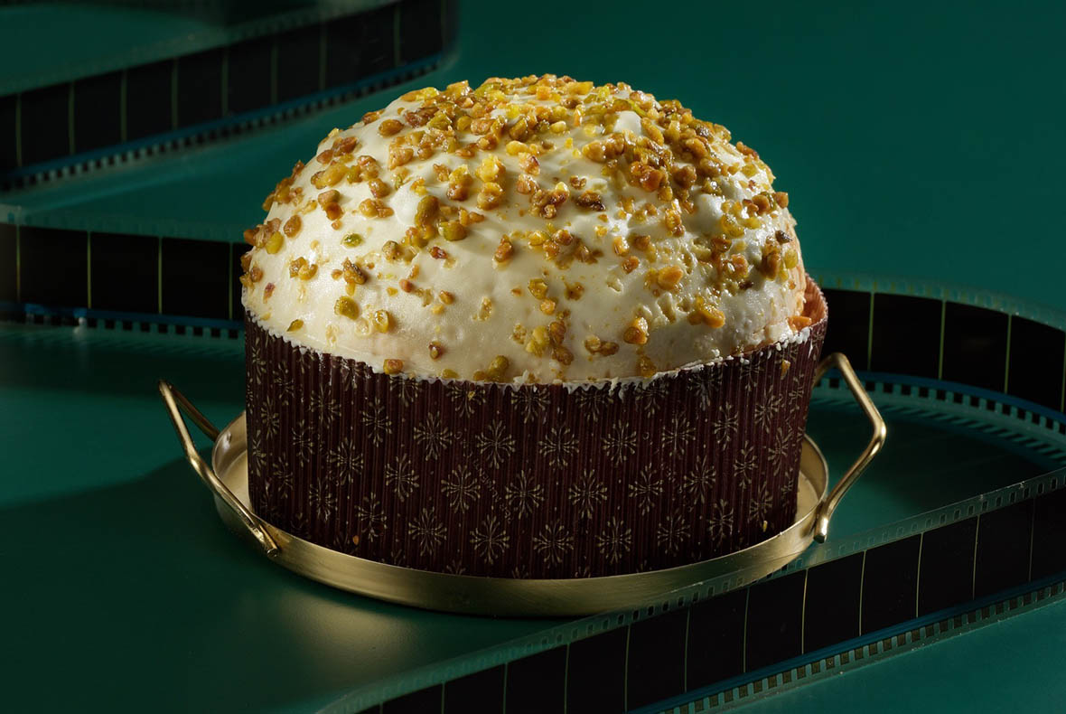 Panettone-style sweet bread with white icing and chopped pistachios in patterned paper case on brass serving tray