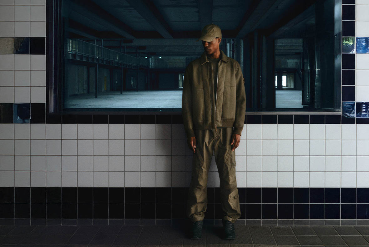 Man in olive-green jacket, cargo trousers and cap standing against tiled wall with large window showing empty industrial space