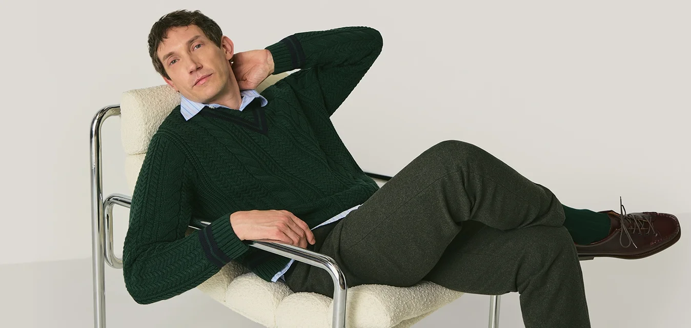Uomo reclinato su una moderna sedia imbottita bianca con struttura in metallo, indossando un maglione verde, pantaloni grigi e scarpe marroni, su uno sfondo semplice.