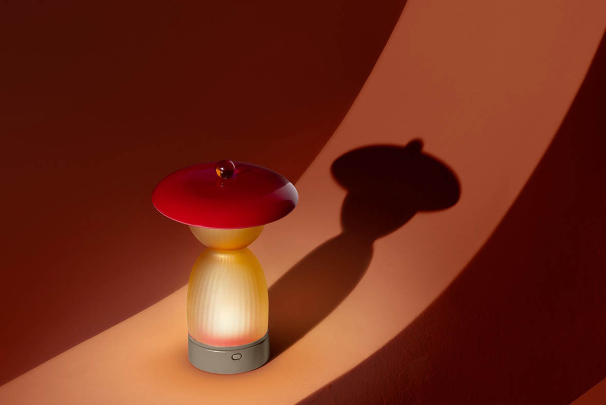 Mushroom-shaped LED lamp with red cap and warm glowing ribbed glass base casting shadow on curved orange background