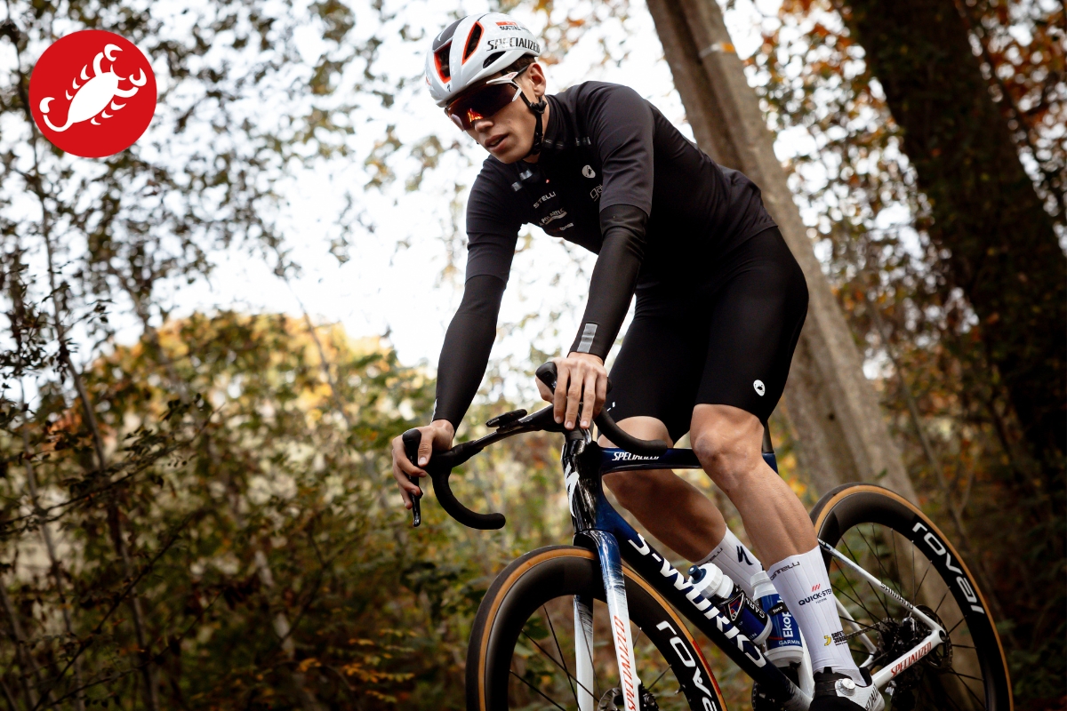 Cyclist in black kit and white helmet riding a road bike through autumn woodland, leaning into a turn and wearing sunglasses