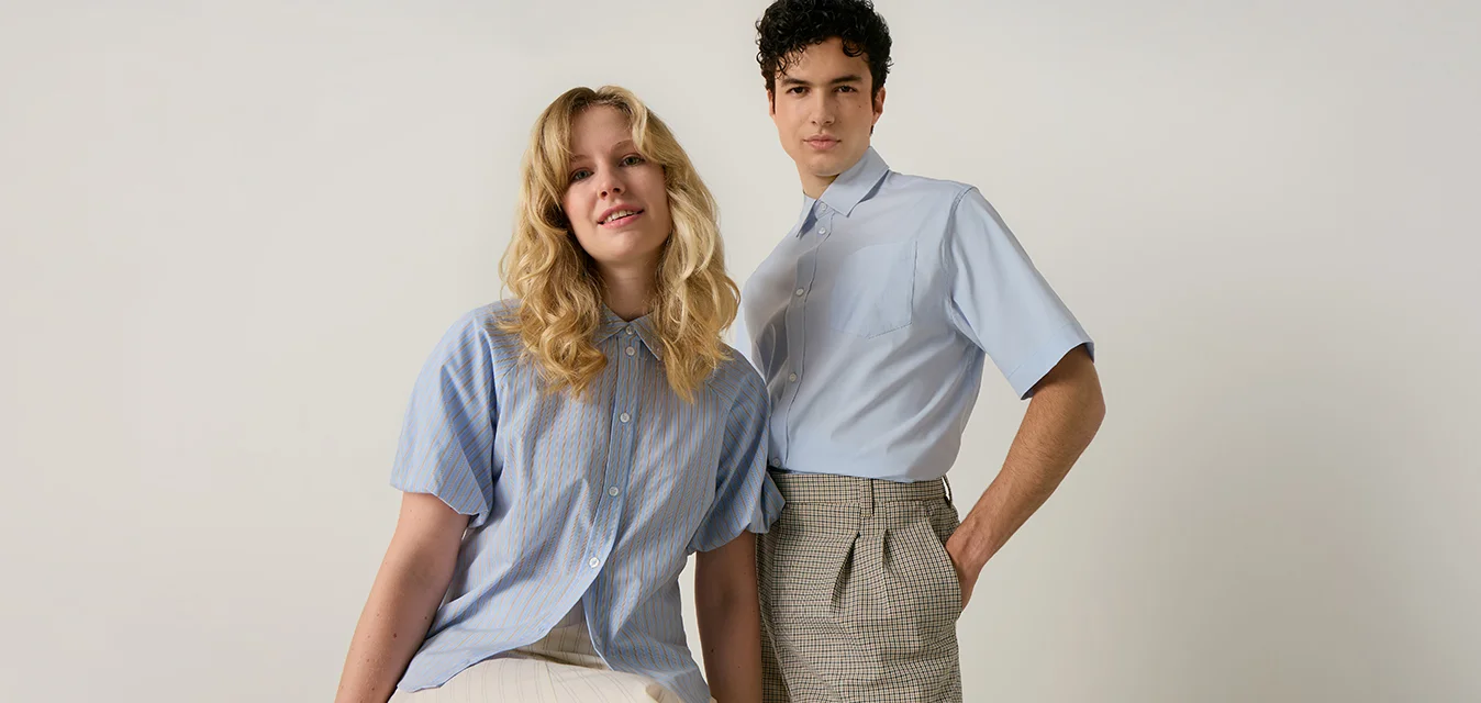 Due giovani adulti che indossano camicie azzurro chiaro, uno seduto con gonna bianca, l'altro in piedi con pantaloni a quadri su uno sfondo semplice.
