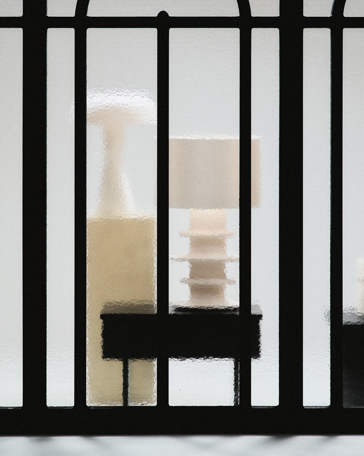 Frosted glass window with black metal grille, blurred silhouettes of table lamps and interior furniture visible through textured glass