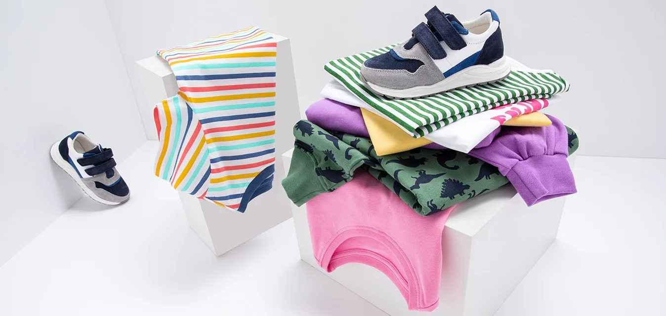 Sneakers per bambini in blu scuro, grigio e bianco, insieme a vestiti piegati: magliette a righe, verdi con stampa di dinosauri, viola, rosa, gialle e bianche.