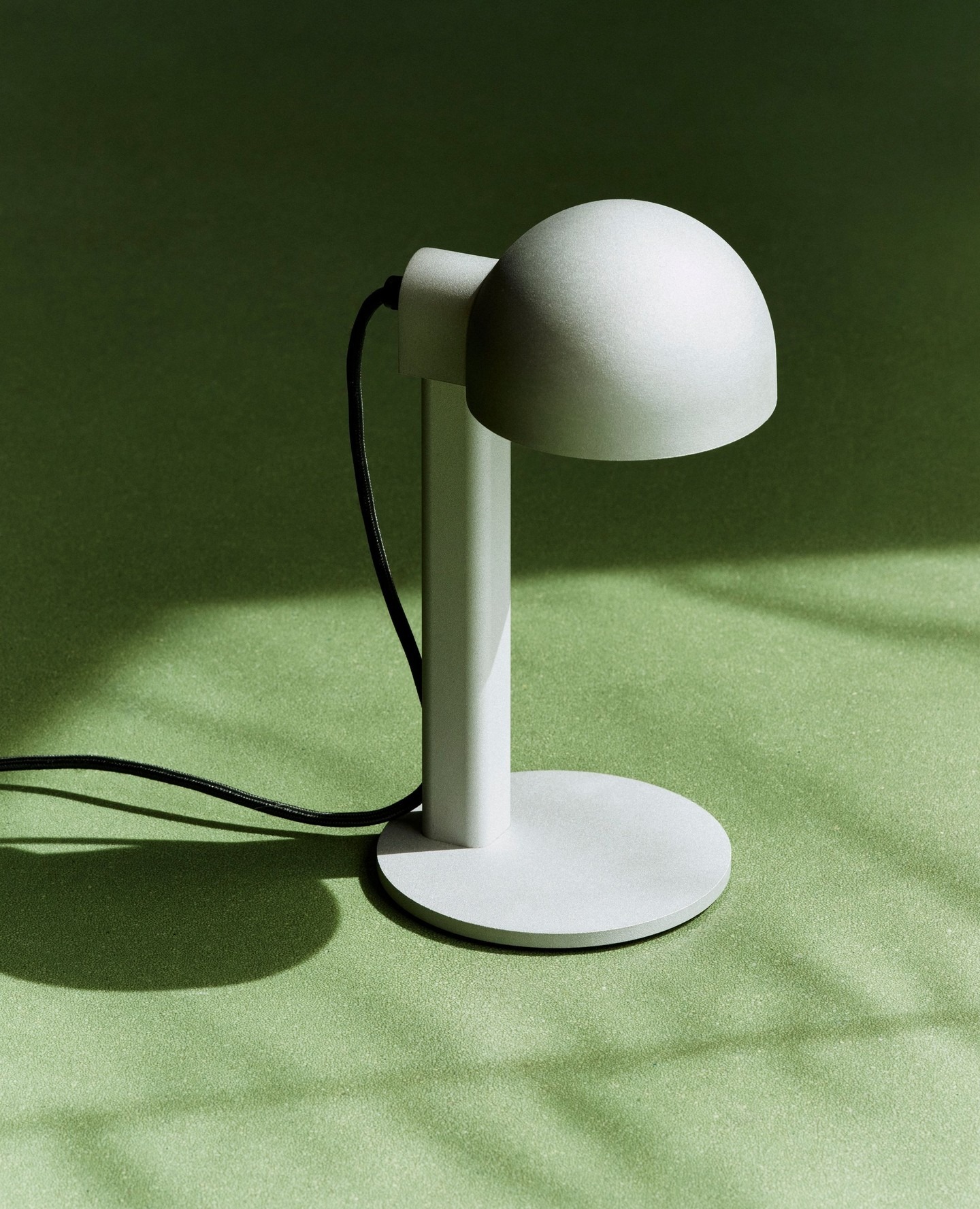 White minimalist table lamp with dome shade and black cord on a green textured surface, casting a soft shadow