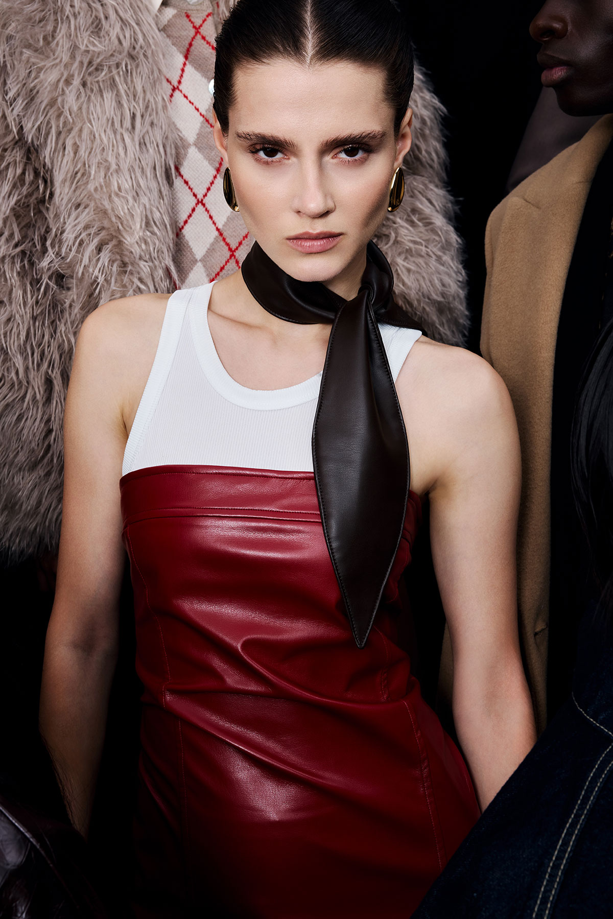 Female model in red leather dress over a white tank, black leather neck scarf and gold hoop earrings, flanked by fur and camel coats